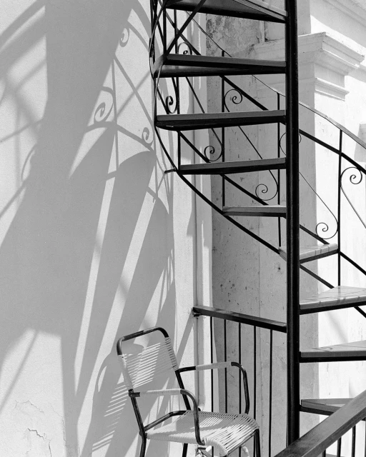 Black and white photo of a metal spiral staircase winding around a column next to a white woven chair, with shadows cast on a white wall.