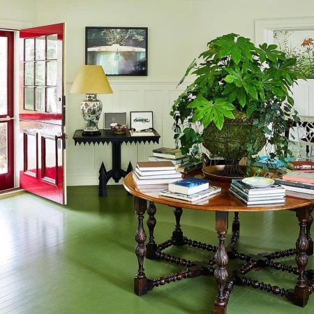 Interior view of a room with a round wooden table topped with books and a plant, complemented by a vintage lamp and green flooring.