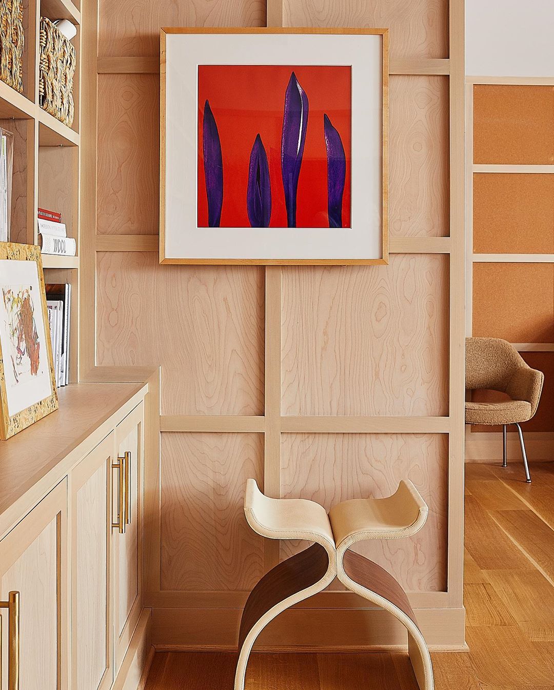 Interior view of a home office nook featuring abstract art, light-colored wood paneled walls, minimalist wooden stool, and hardwood floors.