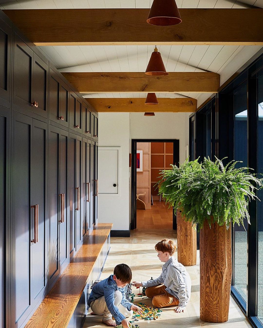 Two young boys play on a wood floor in a hallway with dark blue cabinets, wood benches, pendant lights, and potted ferns.