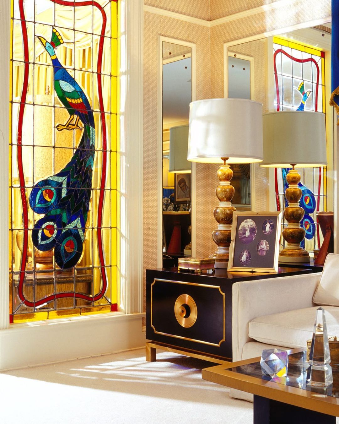 Ornate interior with peacock stained glass window, table lamps on black and gold cabinet, and glass prisms on a coffee table.