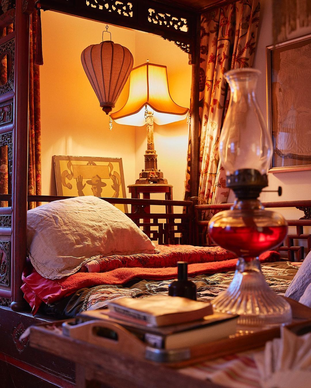 Ornate four-poster bed with a patterned duvet and pillow is visible, featuring an oil lamp and various other antique objects.