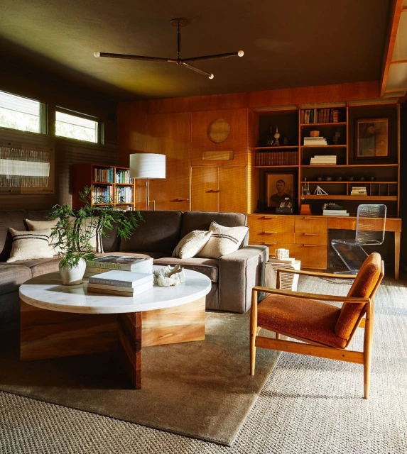 Living room interior showcases a brown sectional sofa, round white coffee table, and orange armchair, complemented by wood paneling and bookshelves.