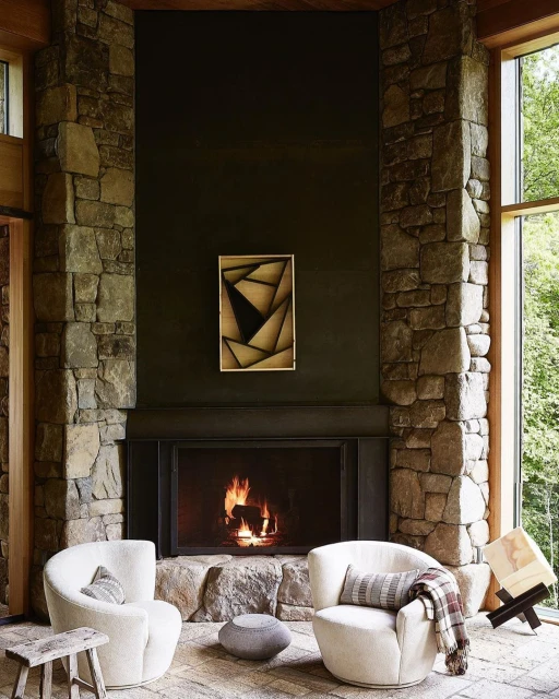 Interior view of a stone fireplace with a burning fire, two white armchairs, an abstract painting, and stone walls and floors.