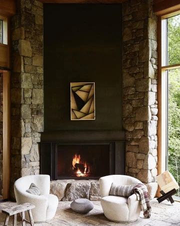 Interior view of a stone fireplace with a burning fire, two white armchairs, an abstract painting, and stone walls and floors.