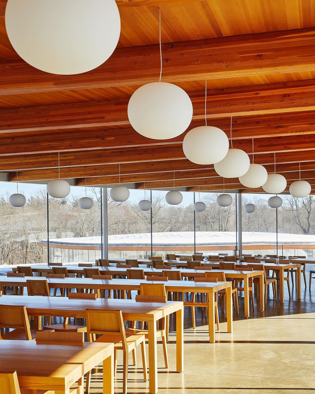 Interior of a modern cafeteria or large dining hall with rows of wooden tables and chairs, illuminated by globe pendant lights.