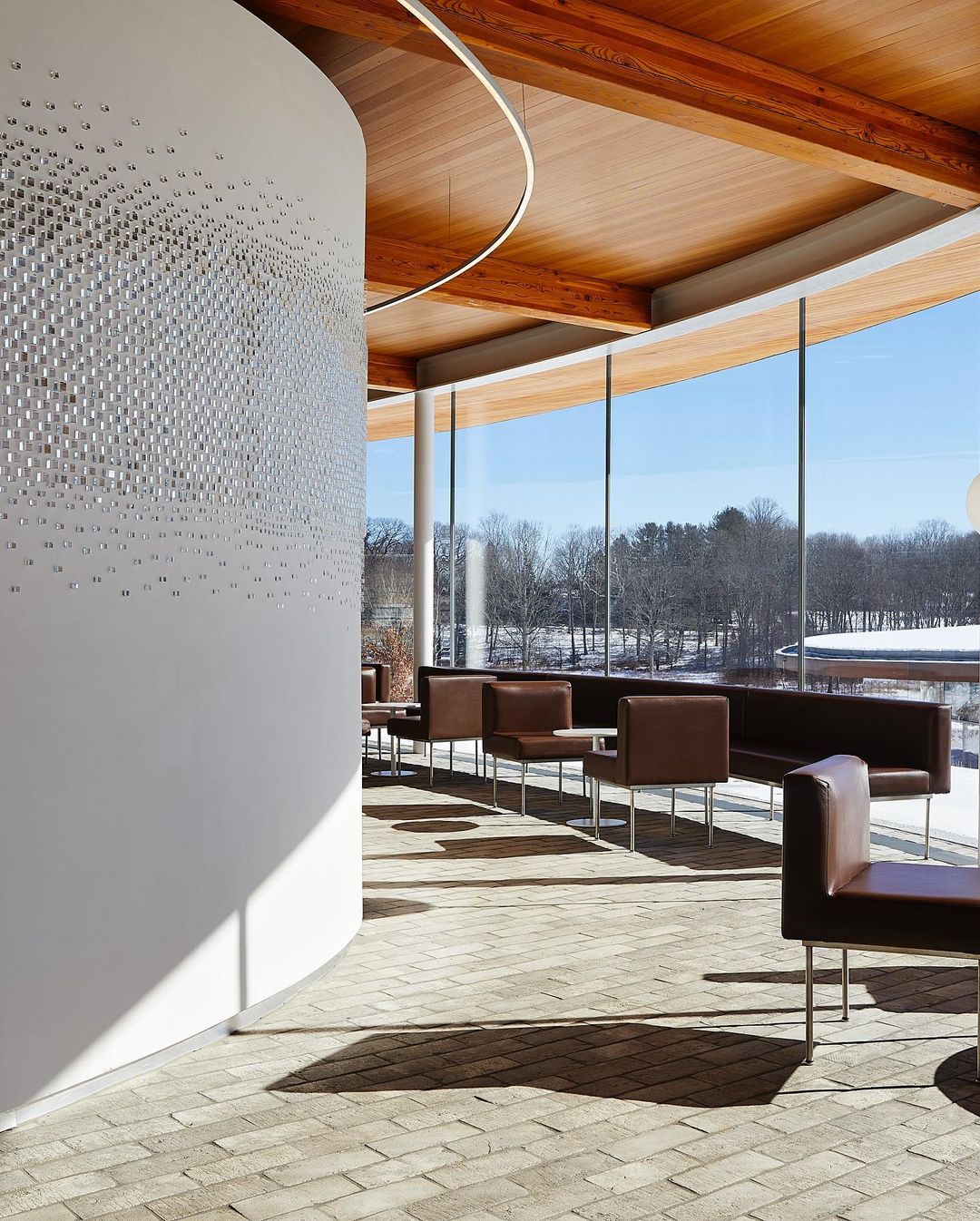 Architectural interior of a modern lounge with brown leather chairs and sofas, white walls with textured design, brick floor, and large windows.
