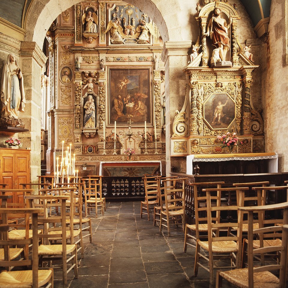 Interior view of a church sanctuary with rows of wooden chairs, ornate carved altars, religious artwork, and stone archways.