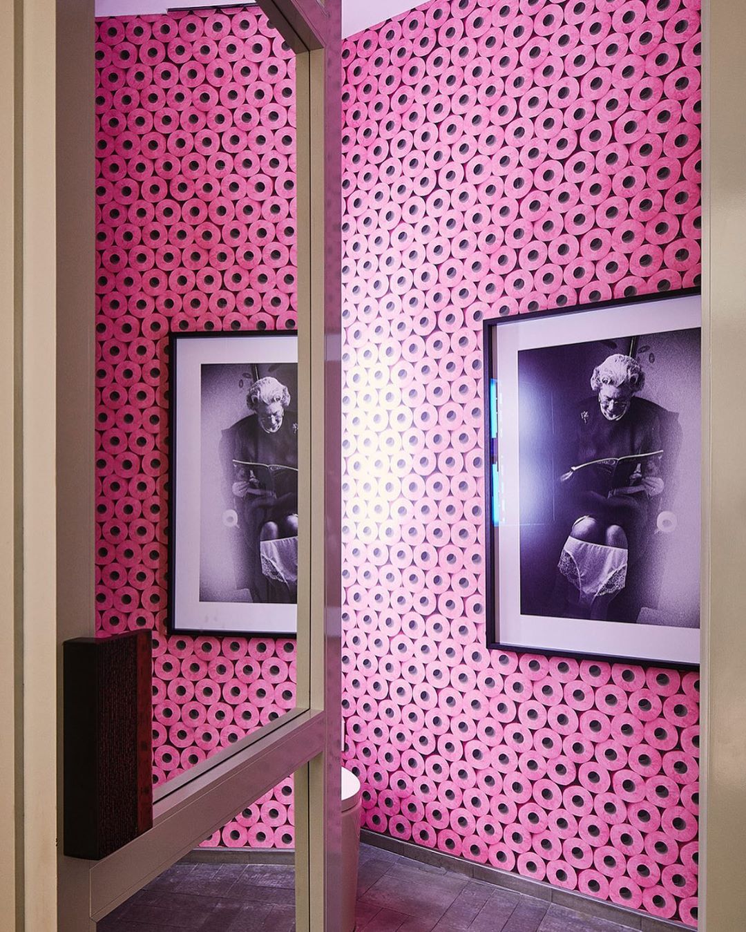 Pink-themed bathroom features walls covered in toilet paper roll wallpaper, framed monochrome art, and a glimpse of a toilet bowl.