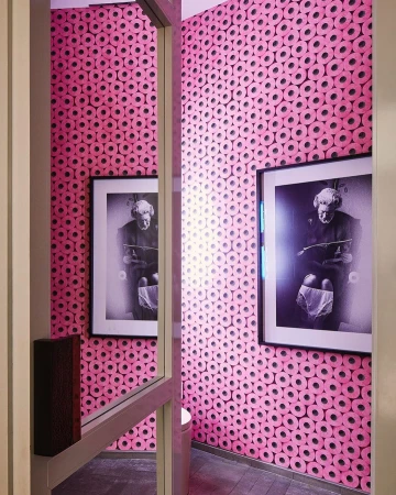 Pink-themed bathroom features walls covered in toilet paper roll wallpaper, framed monochrome art, and a glimpse of a toilet bowl.