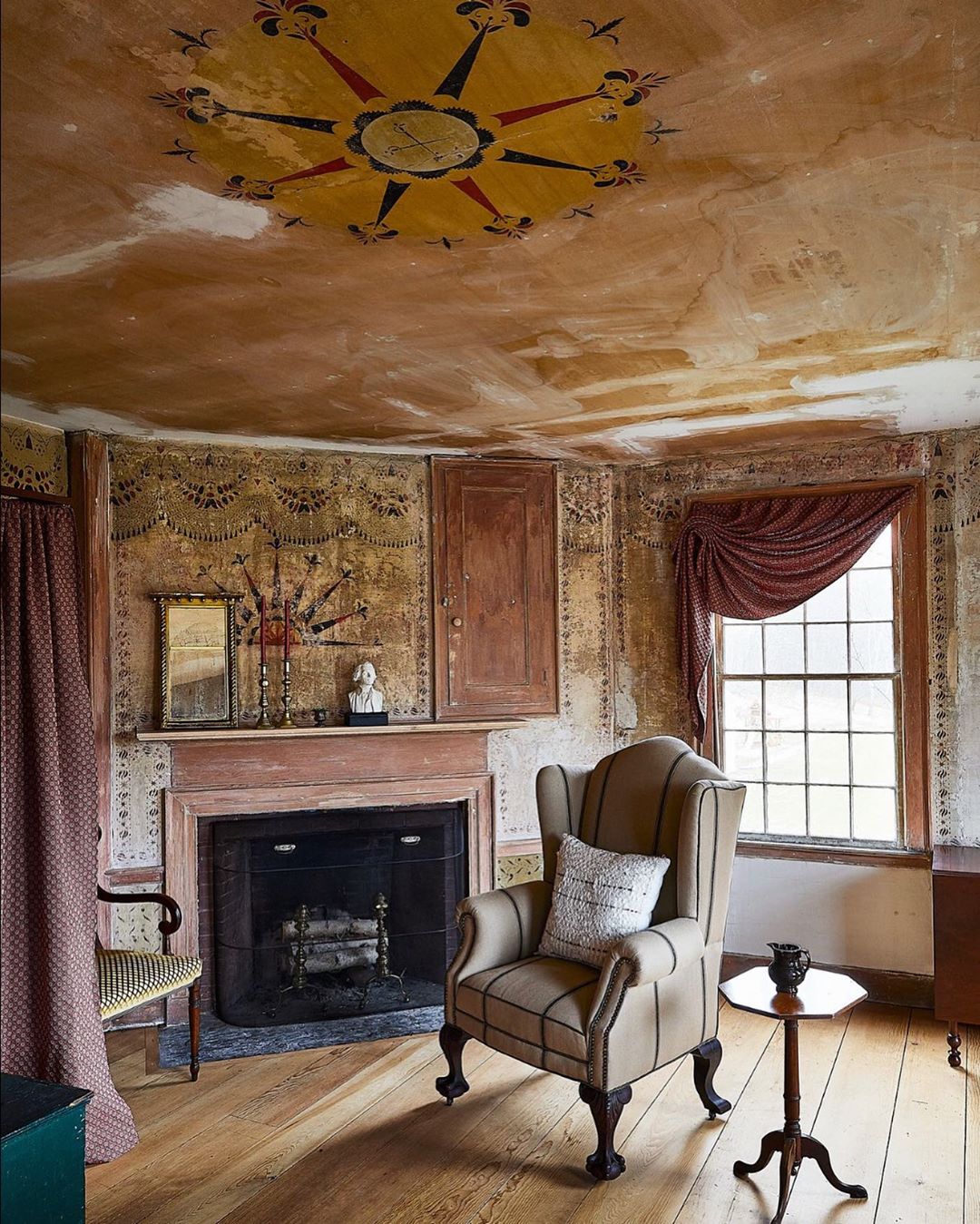 Interior of an old-fashioned room with a fireplace, painted walls and ceiling, a patterned chair, wooden floors, and a window.