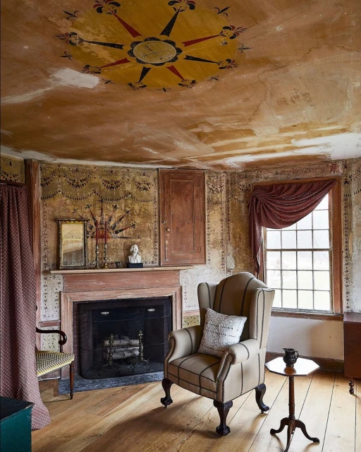 Interior of an old-fashioned room with a fireplace, painted walls and ceiling, a patterned chair, wooden floors, and a window.