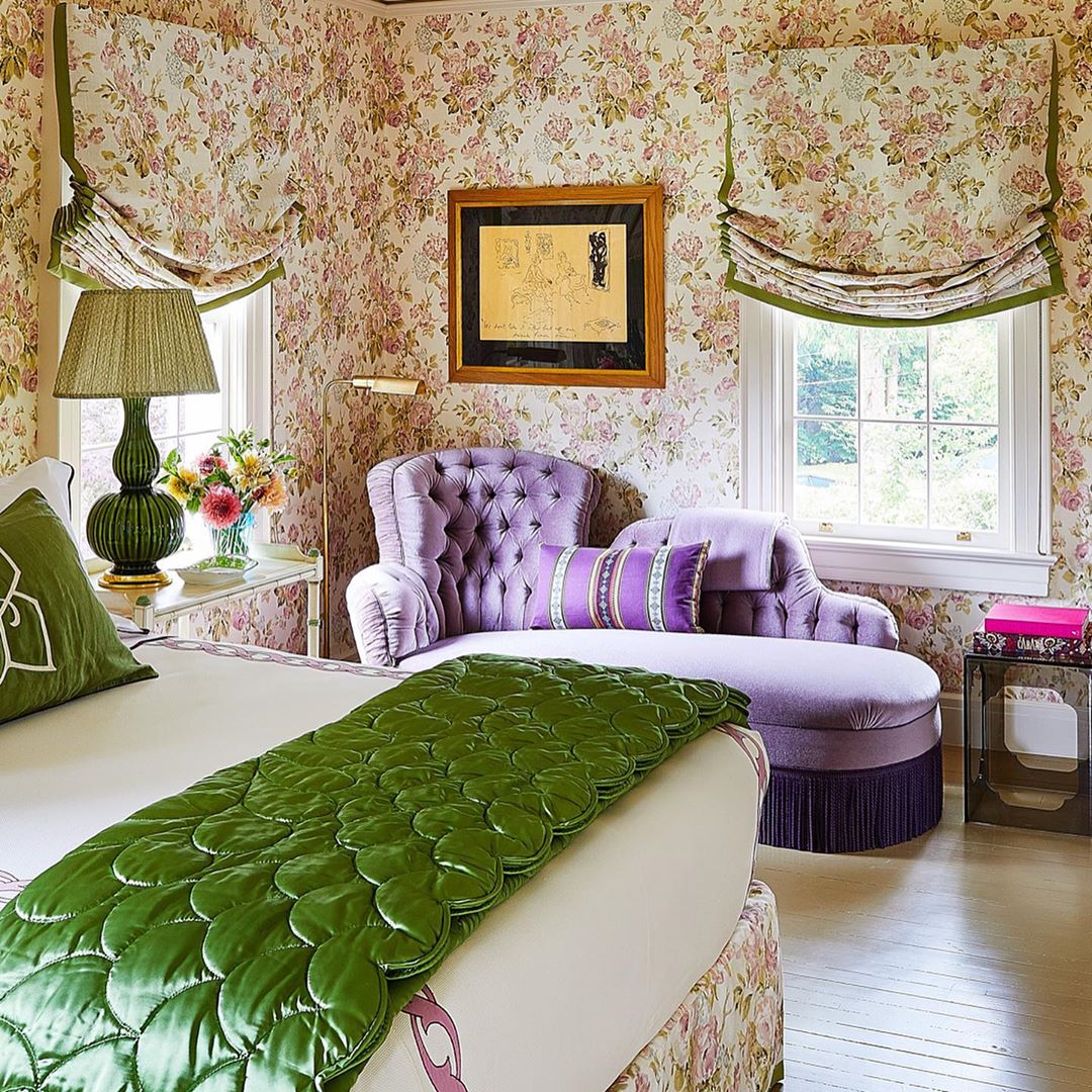 Interior bedroom with floral wallpaper, a purple chaise lounge, green decorative pillows and bedspread, and a window with green trim.