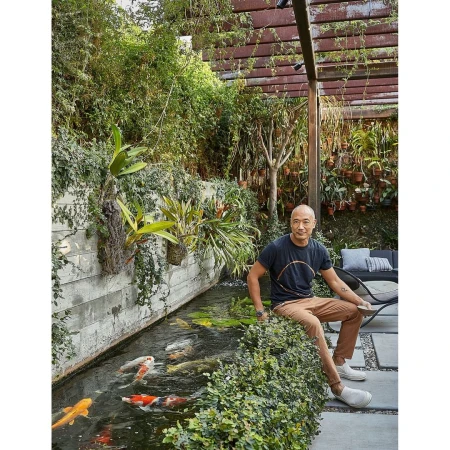 Man seated near a pond with colorful koi fish and lush greenery, under a pergola in an outdoor setting on a sunny day.