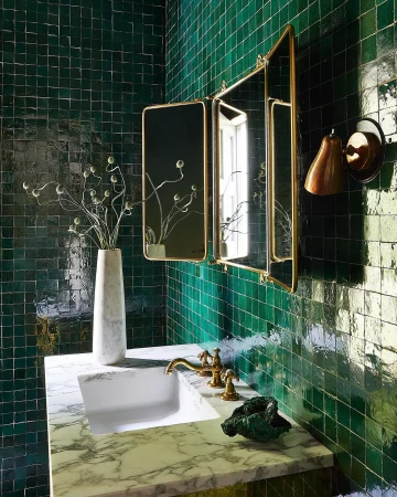 Bathroom interior featuring a marble countertop, white sink, brass fixtures, a trifold mirror, and green tiled walls in a home setting.