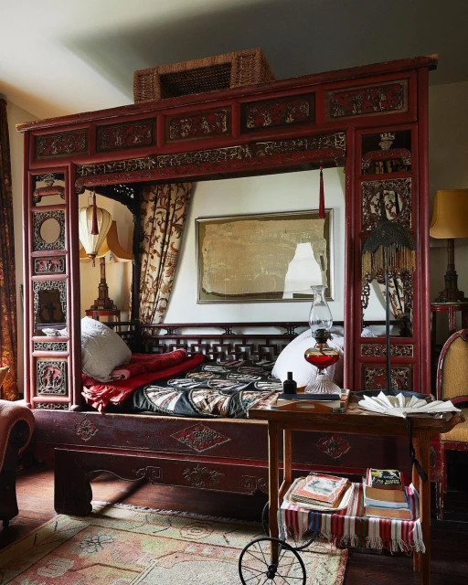 Ornate canopy bed in an indoor setting with decorative objects, a table, and a patterned rug adding to the room's detailed interior.