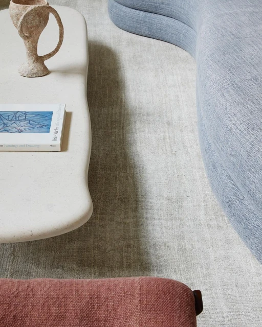 Interior shot of a cream-colored travertine coffee table with a vase and book, a blue couch, and a pink ottoman on a gray carpet.