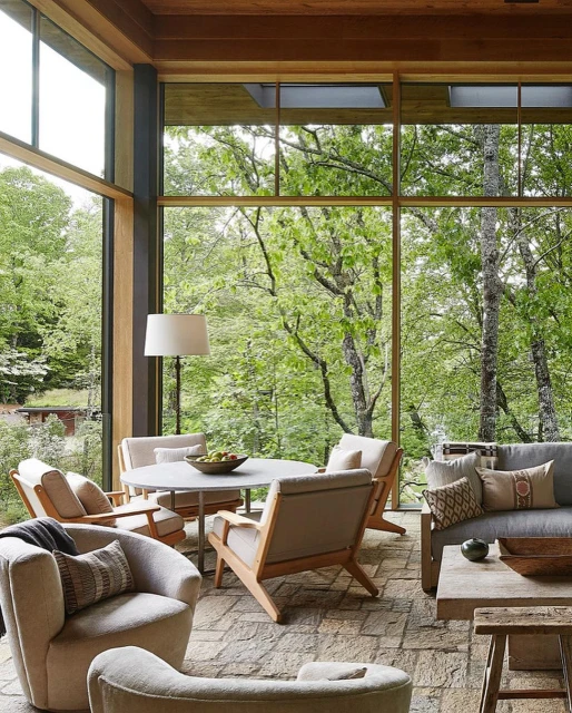 Living room interior with neutral-tone chairs and sofa arranged around a round table, with large windows showing a lush green forest backdrop.