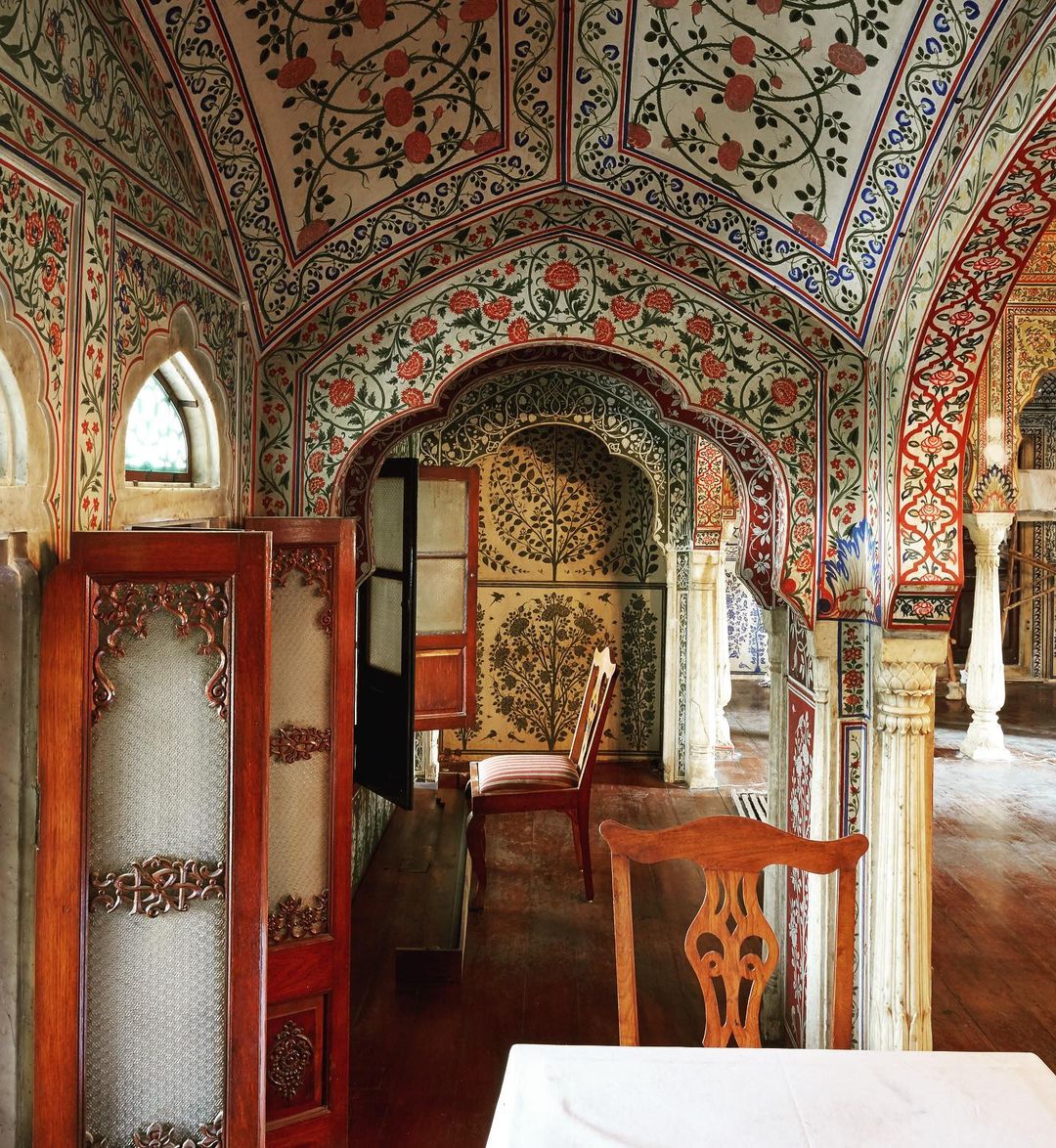 Ornate Indian-style interior featuring red floral motifs, arches, wooden screens, and a table with white tablecloth in muted light.