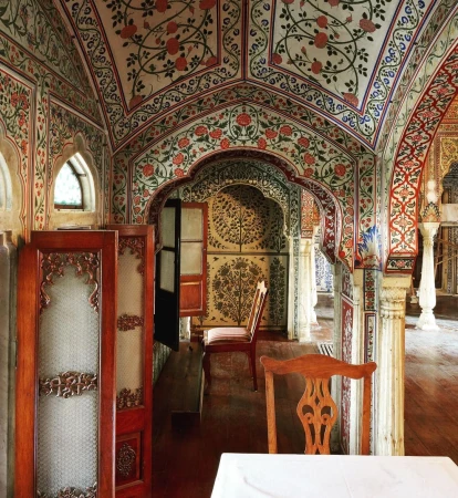 Ornate Indian-style interior featuring red floral motifs, arches, wooden screens, and a table with white tablecloth in muted light.