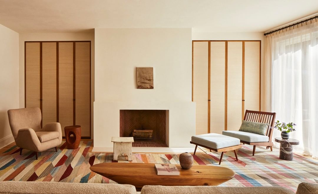 Living room interior features a fireplace, armchairs, and a colorful rug, creating a cozy and inviting atmosphere with natural light.