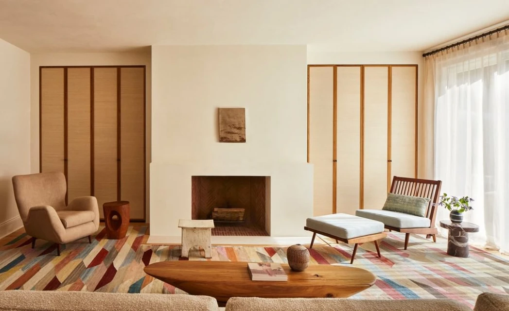 Living room interior features a fireplace, armchairs, and a colorful rug, creating a cozy and inviting atmosphere with natural light.