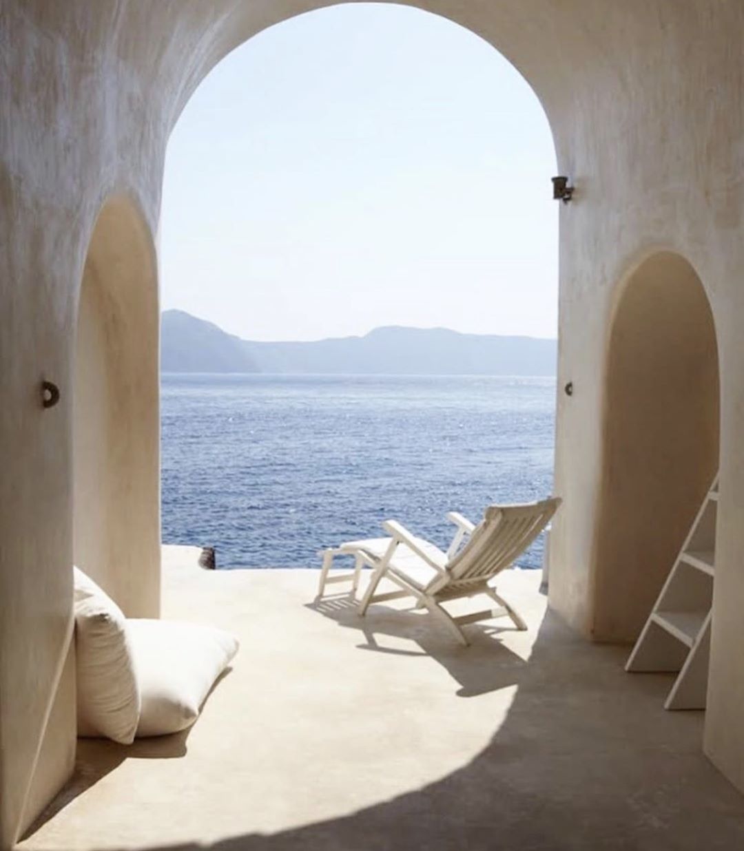 Interior view through an arched opening showcasing a white deck chair on a patio with a sea view, complemented by white cushions and ladder.