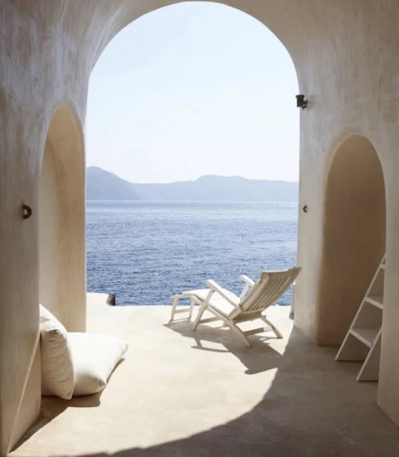 Interior view through an arched opening showcasing a white deck chair on a patio with a sea view, complemented by white cushions and ladder.