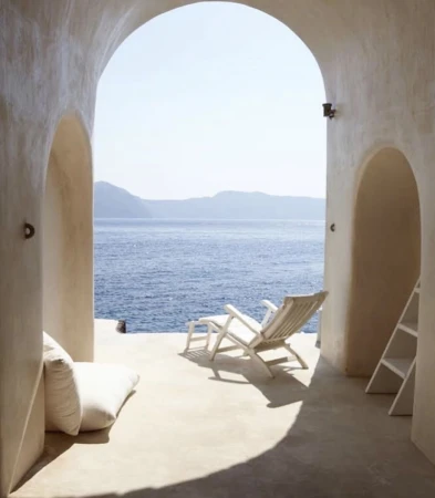 Interior view through an arched opening showcasing a white deck chair on a patio with a sea view, complemented by white cushions and ladder.