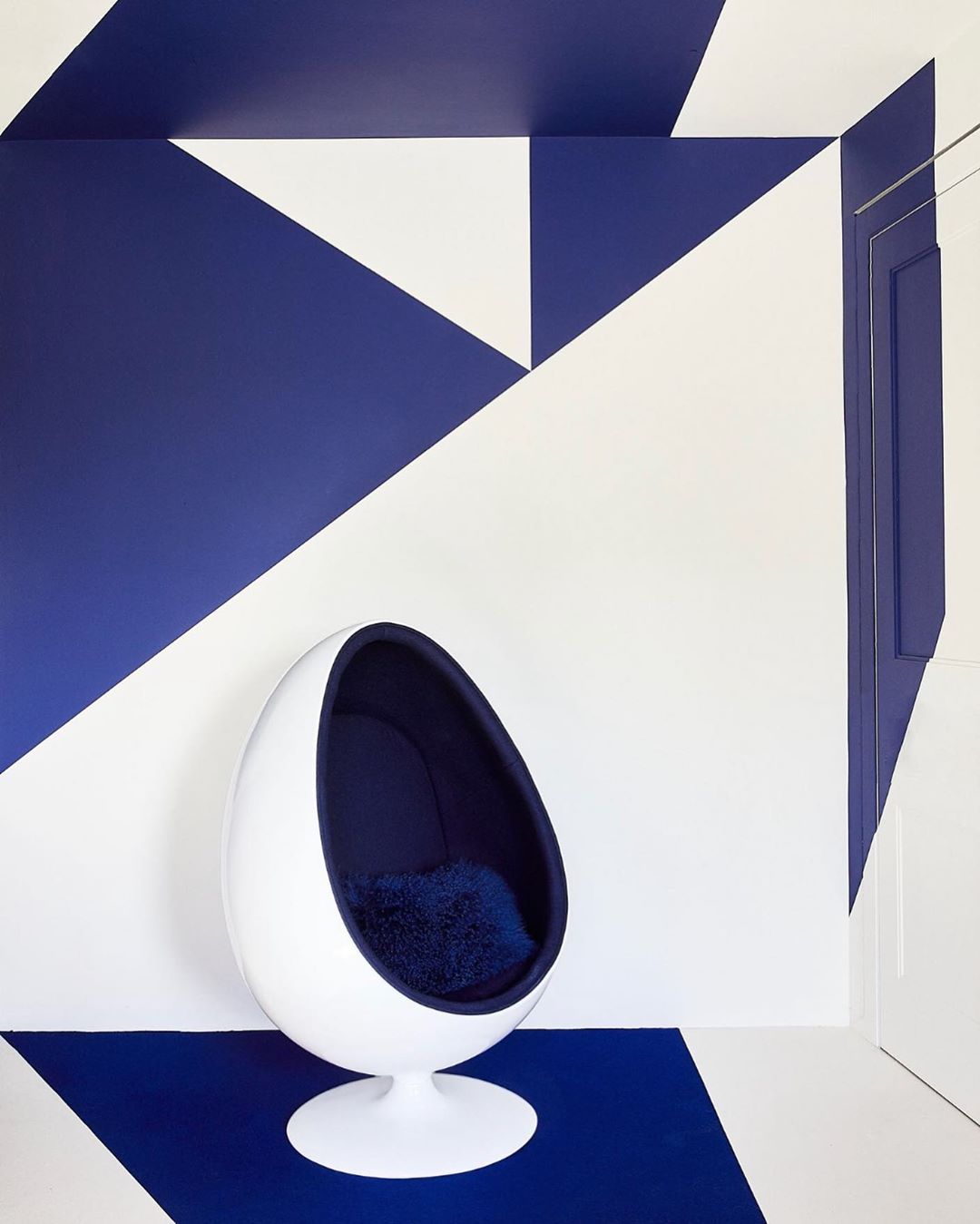 Interior shot of a white egg chair with blue upholstery on a blue and white triangular patterned floor and wall.