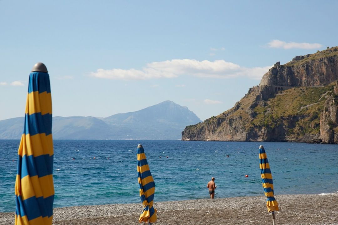 Scenic beach view featuring blue and yellow striped umbrellas on a pebble beach, calm blue sea, a swimmer, and a rocky hillside backdrop.