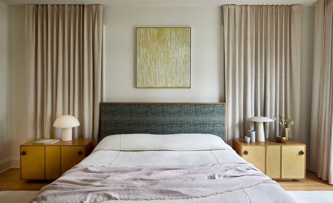 Bedroom interior featuring a made bed with a patterned headboard, bedside tables with lamps, and neutral colored curtains in a home setting.