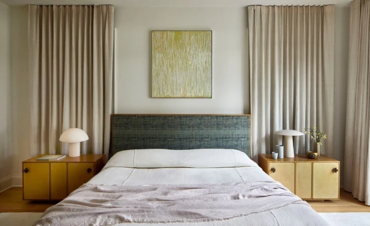 Bedroom interior featuring a made bed with a patterned headboard, bedside tables with lamps, and neutral colored curtains in a home setting.