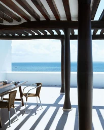 Sunny terrace with dark wooden beams, round columns, and a dining table overlooking the blue ocean by William Abranowicz
