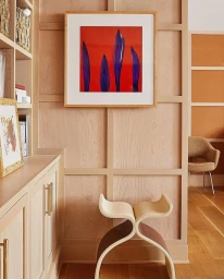 Framed botanical art print with purple pods on red background mounted on pale wood paneling wall above a curved plywood stool