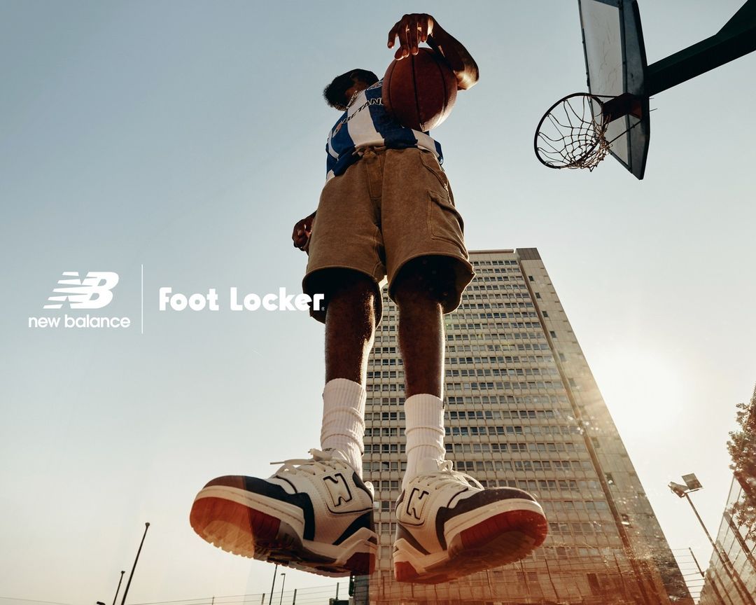 A low-angle shot captures a basketball player mid-air, ball in hand, against a backdrop of a building and blue sky with New Balance and Foot Locker logos.