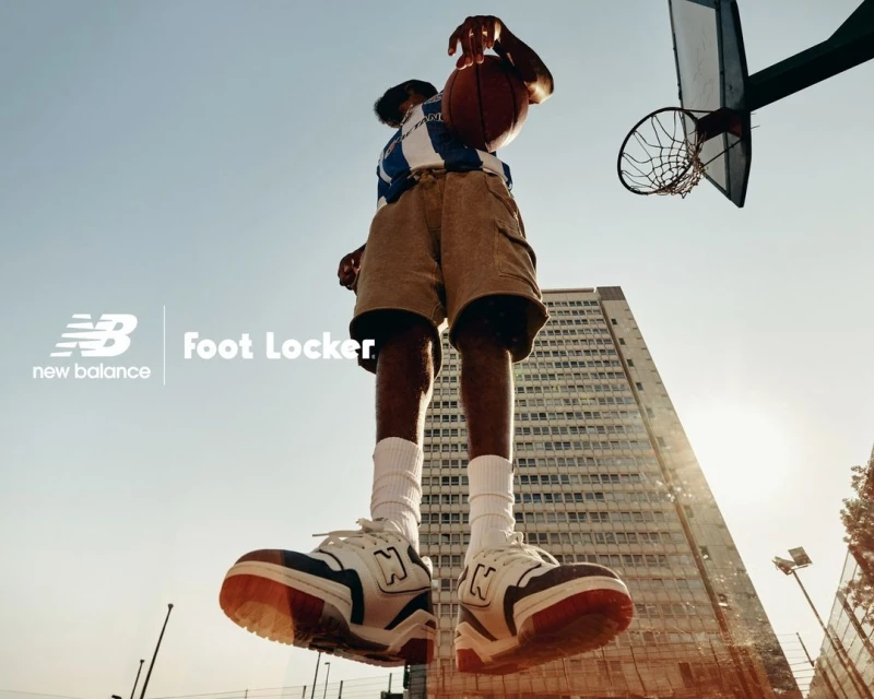 A low-angle shot captures a basketball player mid-air, ball in hand, against a backdrop of a building and blue sky with New Balance and Foot Locker logos.