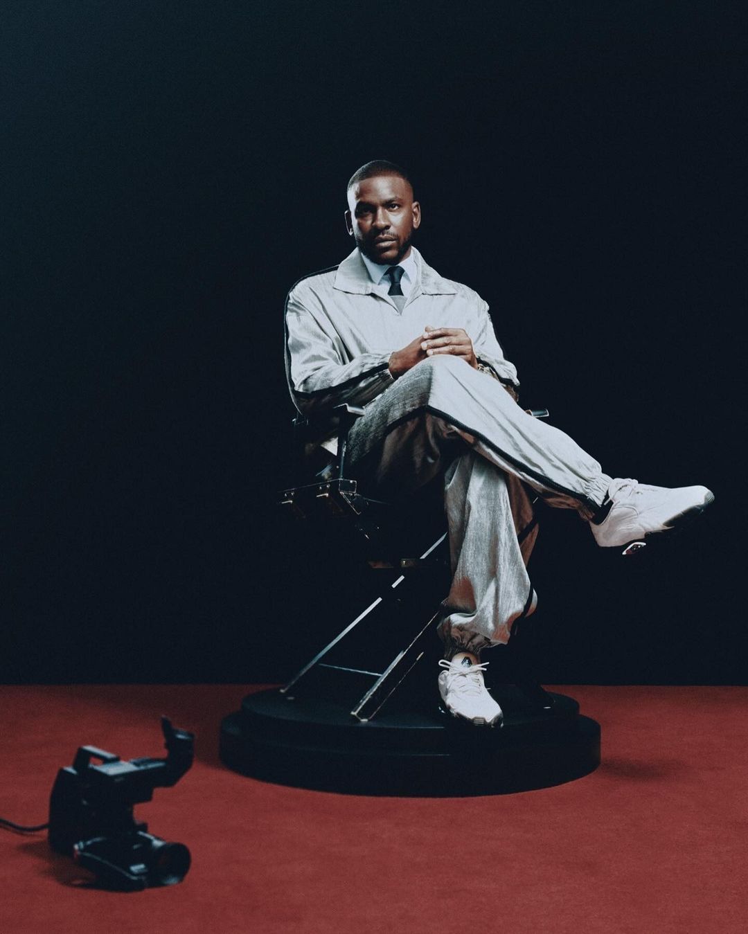 Studio portrait of a stylish adult male seated in a director's chair, wearing a light grey tracksuit, dress shirt, and tie, against a dark backdrop.