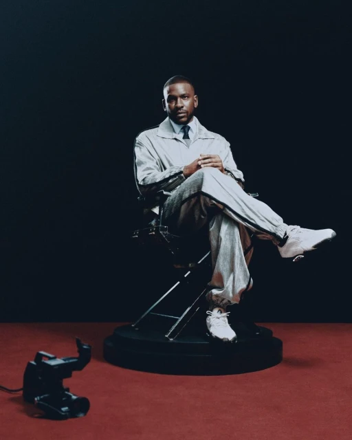 Studio portrait of a stylish adult male seated in a director's chair, wearing a light grey tracksuit, dress shirt, and tie, against a dark backdrop.