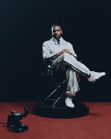 Studio portrait of a stylish adult male seated in a director's chair, wearing a light grey tracksuit, dress shirt, and tie, against a dark backdrop.