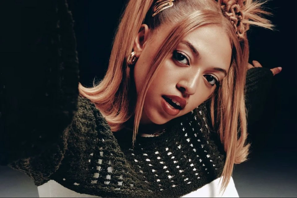 Portrait of a young adult female with auburn hair styled in space buns, wearing a crocheted top against a dark backdrop.