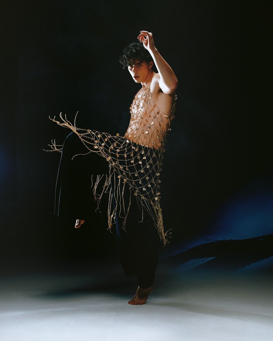 A stylish young adult male wearing a gold decorative net, stands in a dimly lit studio with a blue gradient in the background.
