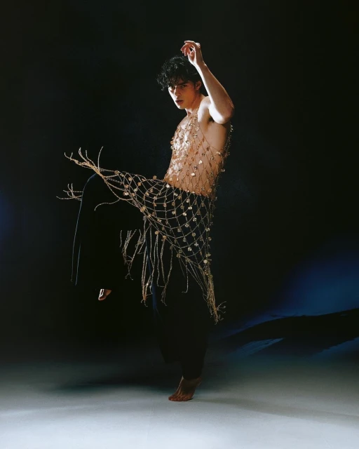A stylish young adult male wearing a gold decorative net, stands in a dimly lit studio with a blue gradient in the background.