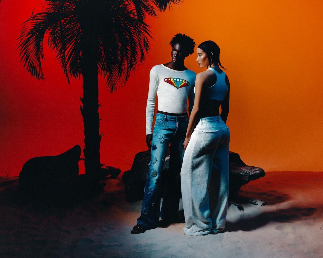 Two models posing in a studio set with a red and orange backdrop and a silhouetted palm tree by William Arcand