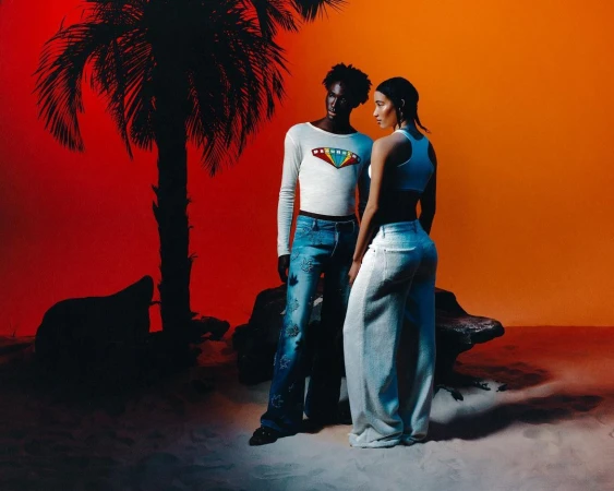 Two models posing in a studio set with a red and orange backdrop and a silhouetted palm tree by William Arcand