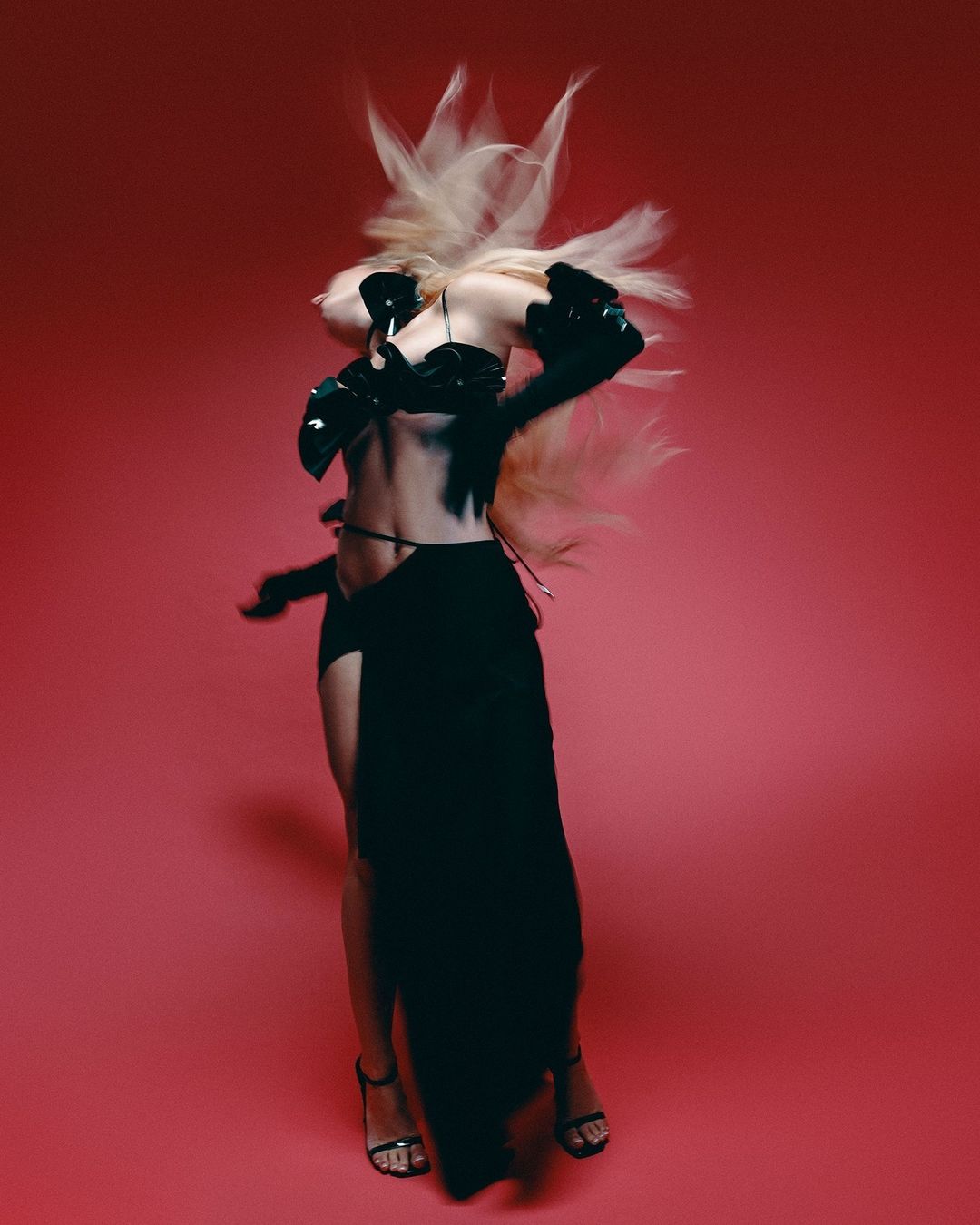 A woman with blonde hair dances in a black outfit against a vibrant pink backdrop, showcasing dynamic movement and modern fashion.