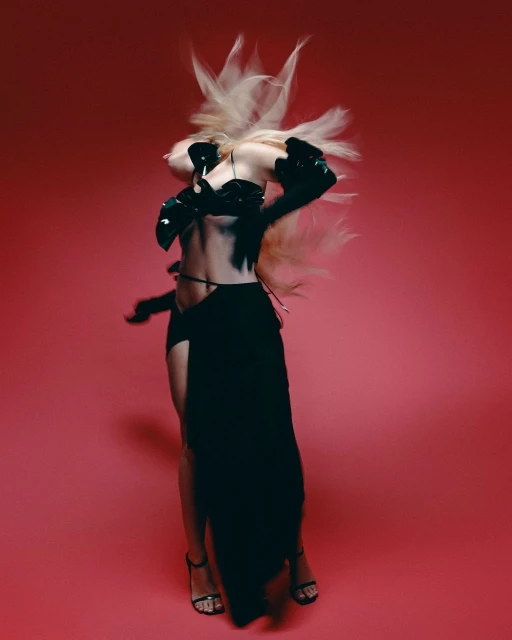 A woman with blonde hair dances in a black outfit against a vibrant pink backdrop, showcasing dynamic movement and modern fashion.