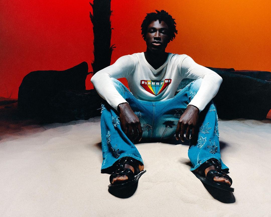 A dark-skinned young adult sits in a stylish casual outfit. He wears a white logo tee, light blue palm-print jeans, and black platform sandals.