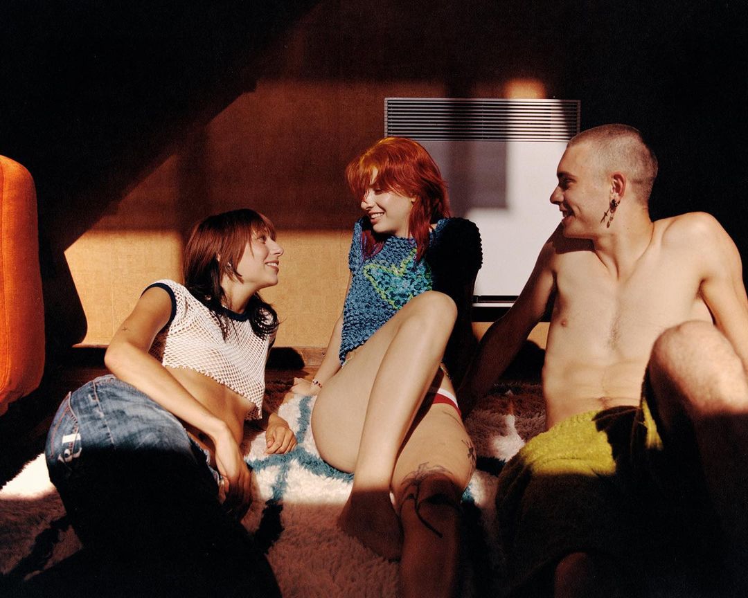 Three people are sitting on a carpet indoors in a brightly lit room. Two women and a man are smiling at each other in natural light.