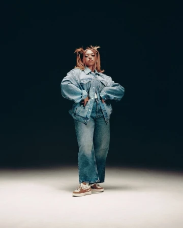Full-length portrait of a person in an oversized denim jacket and matching jeans, posing against a neutral dark gradient background.